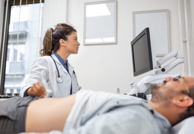Careful female doctor in white coat sitting in front of an ultrasound apparatus and conducting abdominal diagnostics with transducer